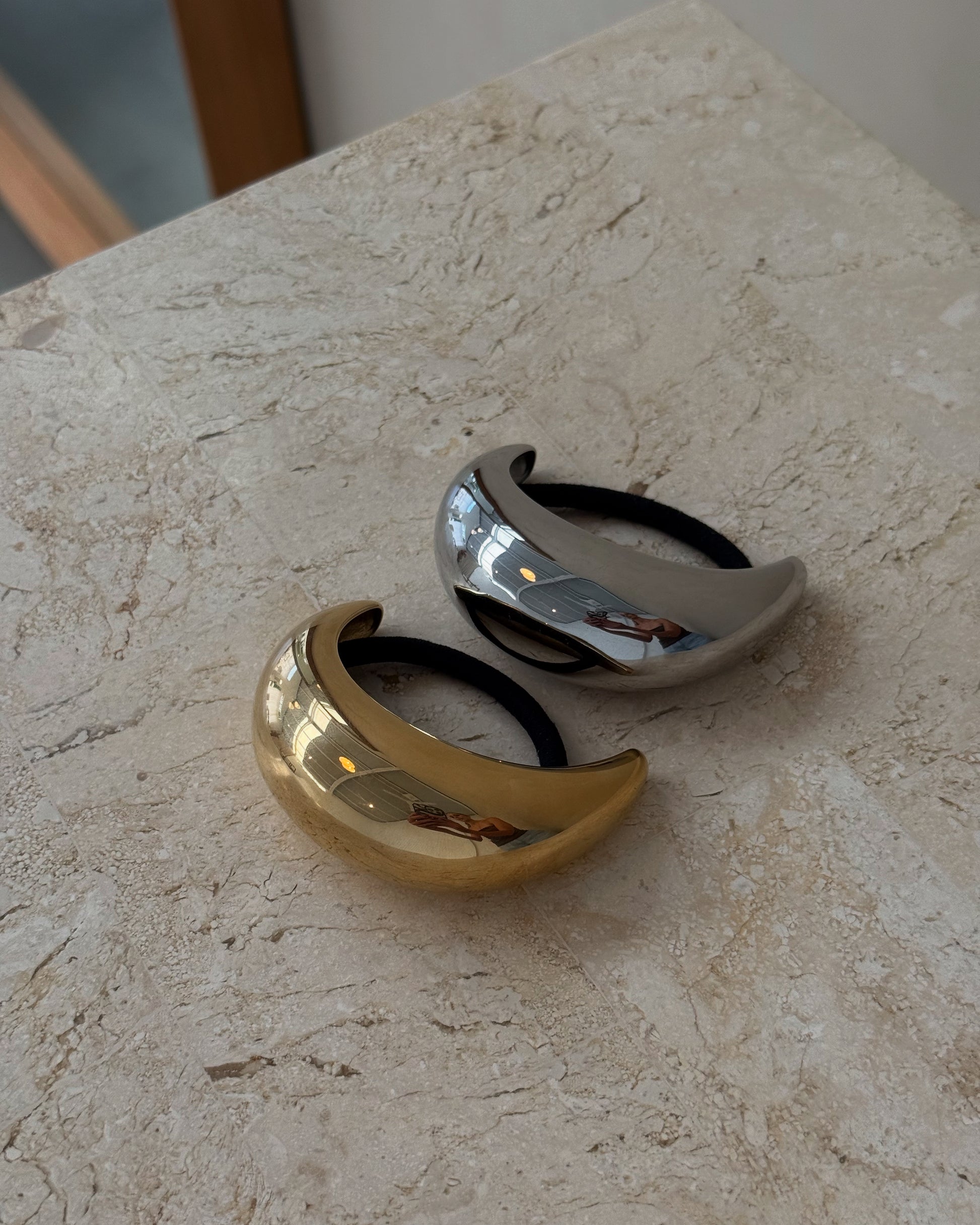 Two cuff hair ties, one in 18k gold-plated and the other in sleek silver finish, displayed on a textured surface.